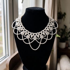 May include: A white pearl collar necklace featuring a multi-layered, intricate lattice design. The piece is constructed from numerous small, round, cream-colored beads arranged in a scalloped, draped pattern that encircles the neck area.