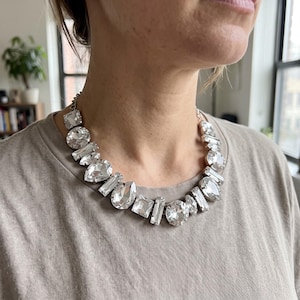 May include: A statement necklace featuring a series of clear, faceted glass crystals in various shapes, including round, pear, square, and rectangular cuts, set in a silver-toned metal frame that rests against the base of the neck.