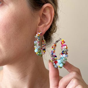 Puede incluir: Un par de pendientes de aro con una densa disposición de cuentas de vidrio multicolores en tonos azul, verde, rojo, amarillo y blanco. Las cuentas varían en tamaño y textura, creando una superficie vibrante sobre el marco metálico circular.