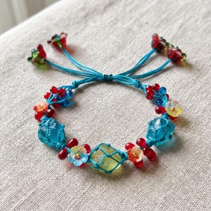 May include: A bracelet featuring a bright blue cord woven with translucent blue and yellow stones, small red beads, and delicate flower-shaped glass beads. The ends of the cord are finished with two small red and green flower-shaped glass beads.