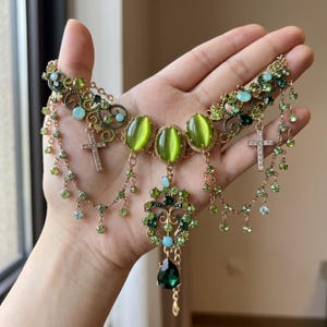 Può includere: Una collana color oro con tre cabochon centrali ovali in occhio di gatto verde, dettagli in filigrana, catenelle pendenti con piccoli cristalli verdi, due ciondoli a croce color argento e un pendente a goccia verde scuro.