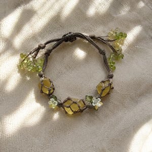 May include: A handmade bracelet featuring yellow gemstones and green beads. The bracelet is crafted with a brown cord and adorned with small, delicate floral accents. The design incorporates natural elements, creating a unique and earthy aesthetic.
