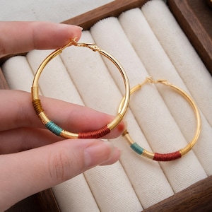 Op de afbeelding: Een paar goudkleurige oorringen met secties omwikkeld met dun draad. De draadaccenten zijn gekleurd in mosterdgeel, teal en donkerrood, afgewisseld met segmenten van de gepolijste goudkleurige metalen ringstructuur.