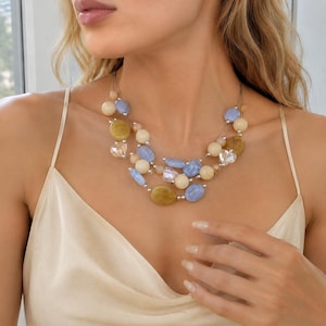 Puede incluir: Un collar de múltiples hilos con un surtido de cuentas lisas y pulidas en tonos azul pálido, crema y amarillo mostaza, intercaladas con pequeños espaciadores de metal plateado y detalles transparentes en forma de corazón sobre hilos finos.
