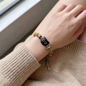 May include: A beaded bracelet featuring a black cat charm. The bracelet has colorful beads in red, yellow, green, and brown, with silver-colored metal accents. A tassel with rainbow-colored threads hangs from the bracelet.