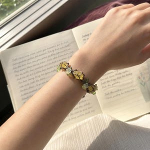 May include: A delicate bracelet featuring yellow gemstone accents and small green floral details. The bracelet is worn on a wrist, with a notebook visible in the background.