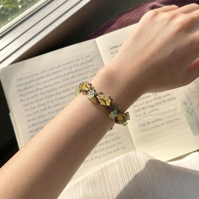 May include: A delicate bracelet featuring yellow gemstone accents and small green floral details. The bracelet is worn on a wrist, with a notebook visible in the background.