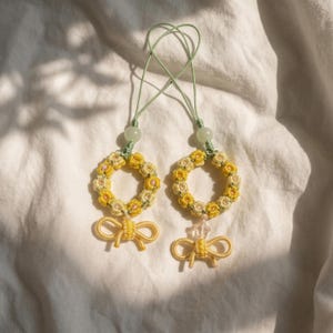 Könnte beinhalten: Zwei handgefertigte gelbe Blumenkranz-Ornamente mit grünen Kordeln und einer gelben Schleife. Jeder Kranz ist mit kleinen gelben Blumen und einer hellgrünen Perle verziert. Die Ornamente werden auf einem weißen Stoffhintergrund präsentiert.