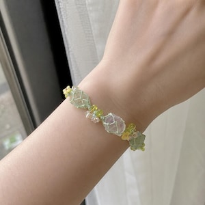May include: A delicate bracelet featuring light green and clear beads, with accents of yellow and small white flower-shaped beads. The bracelet is designed with a floral theme and is worn on a wrist.