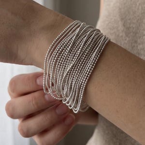 Könnte beinhalten: Ein mehrsträngiges Armband aus zahlreichen dünnen, silberfarbenen Metallketten mit einer texturierten, perlenartigen Oberfläche. Die einzelnen Stränge sind eng beieinander angeordnet und erzeugen eine breite, geschichtete Optik am Handgelenk.