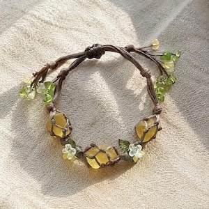 May include: A handmade bracelet featuring yellow gemstones encased in a brown woven macrame design. The bracelet is adorned with small green and clear beads, along with white flower accents. The bracelet is displayed on a neutral-toned surface.
