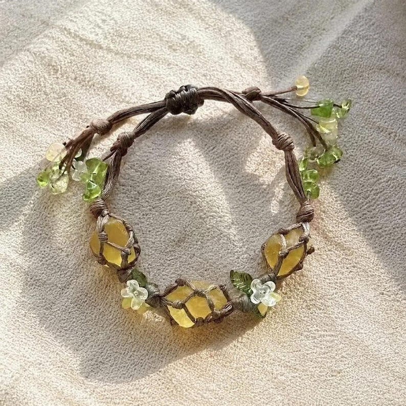 May include: A handmade bracelet featuring yellow gemstones encased in a brown woven macrame design. The bracelet is adorned with small green and clear beads, along with white flower accents. The bracelet is displayed on a neutral-toned surface.