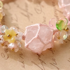 May include: Close-up of a handmade bracelet featuring a large, light pink rose quartz crystal encased in a macrame knot. The bracelet is adorned with small yellow and clear flower beads, and gold accents.