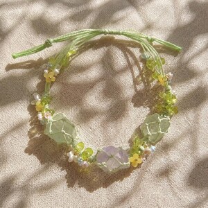 May include: A handmade macrame bracelet featuring light green and purple crystals, accented with small yellow and white floral beads. The bracelet is tied with a light green cord.