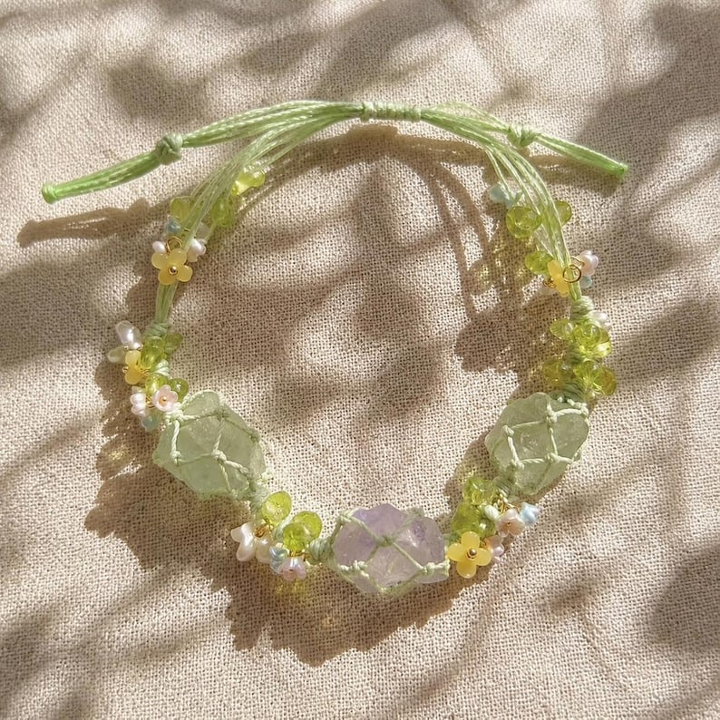 May include: A handmade macrame bracelet featuring light green and purple crystals, accented with small yellow and white floral beads. The bracelet is tied with a light green cord.