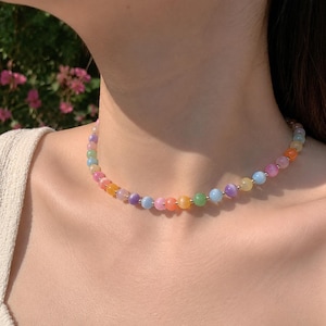 May include: A multi-coloured beaded necklace featuring pastel shades of pink, orange, yellow, green, and blue. The necklace is composed of small, round beads connected by silver-toned metal. The necklace is worn around the neck.