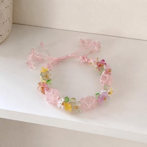 May include: A pink beaded bracelet with a floral design. The bracelet features pink, yellow, green, and purple flowers, along with pink beads. The bracelet is on a white surface.