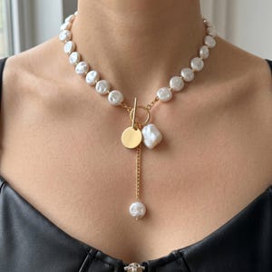 May include: A gold-toned necklace featuring large, irregular white pearls. The necklace has a gold pendant with a small, round charm and a dangling pearl. The necklace is secured with a gold toggle clasp.