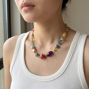 May include: A colorful beaded necklace featuring a variety of shapes and colors. The necklace includes beads in shades of red, pink, purple, blue, and yellow. The clasp is gold-toned.