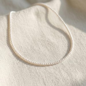 Puede incluir: Delicado collar de perlas compuesto por pequeñas perlas blancas y redondas. El collar se muestra sobre una tela crema texturizada, creando una estética suave y elegante. Las perlas están ensartadas, formando un círculo completo.