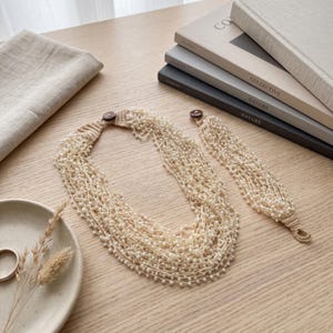 May include: A multi-strand necklace and matching bracelet set featuring numerous small white beads strung on light beige thread. Each piece is finished with a dark brown circular button closure and a crocheted loop, creating a layered appearance.