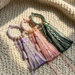 May include: Three handmade tassel ornaments in shades of purple, pink, and green. Each ornament features a braided loop and a long tassel with intricate details. The tassels are adorned with beads and varying braid patterns, resting on a textured, off-white surface.