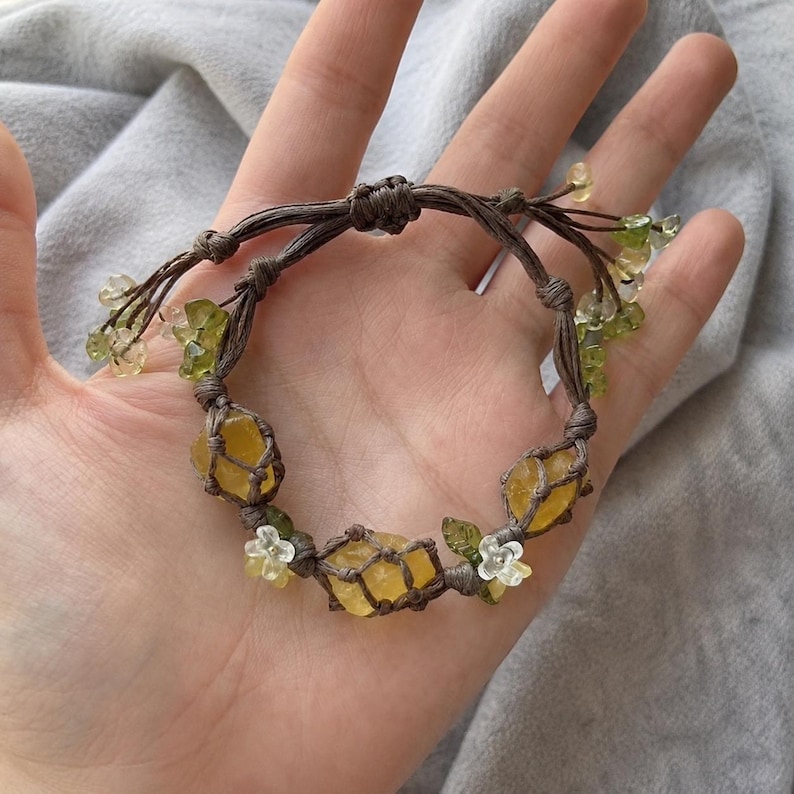 May include: A handmade bracelet featuring yellow and green gemstones, accented with small white flowers and green leaves. The stones are encased in a brown woven macrame design. The bracelet is displayed on a neutral background.
