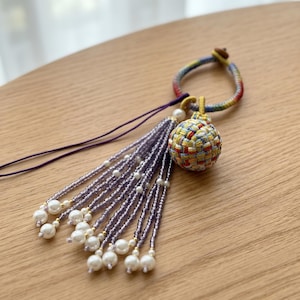 May include: A handcrafted decorative tassel with a woven sphere in yellow, red, blue, and green. The tassel features strands of purple beads and white pearls, attached to a rainbow-colored loop.