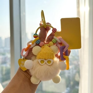 May include: A whimsical keychain featuring a fluffy, cream-colored creature wearing a yellow knit hat and sunglasses. The keychain includes colorful beads, a yellow tag, and a chain with pink, purple, and yellow links. The overall design is playful and fun.