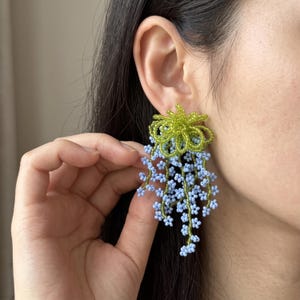 Könnte beinhalten: Ein Perlenohrring mit einem Bündel leuchtend grüner Schlaufen am oberen Ende und mehreren herabhängenden Strängen aus kleinen, hellblauen, blütenförmigen Perlen, die ein florales Design mit einem lebendigen Farbkontrast bilden.
