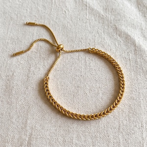 May include: A gold-toned bracelet featuring a woven wheat chain design. The bracelet includes an adjustable sliding bead mechanism with two thin chain extensions ending in small, cylindrical metal tips for a secure and customizable fit.