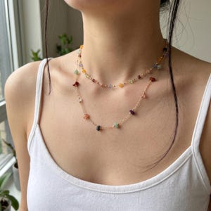 May include: A layered necklace featuring two delicate gold-toned chains adorned with small, multi-coloured, irregularly shaped gemstone beads. The beads are spaced evenly along the chains, showcasing a variety of hues including red, green, blue, and yellow.