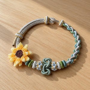 May include: A circular bracelet crafted from braided cord in shades of sage green, light blue, and silver, featuring a prominent three-dimensional yellow sunflower charm with a dark brown center and intricate decorative knots with gold thread accents.