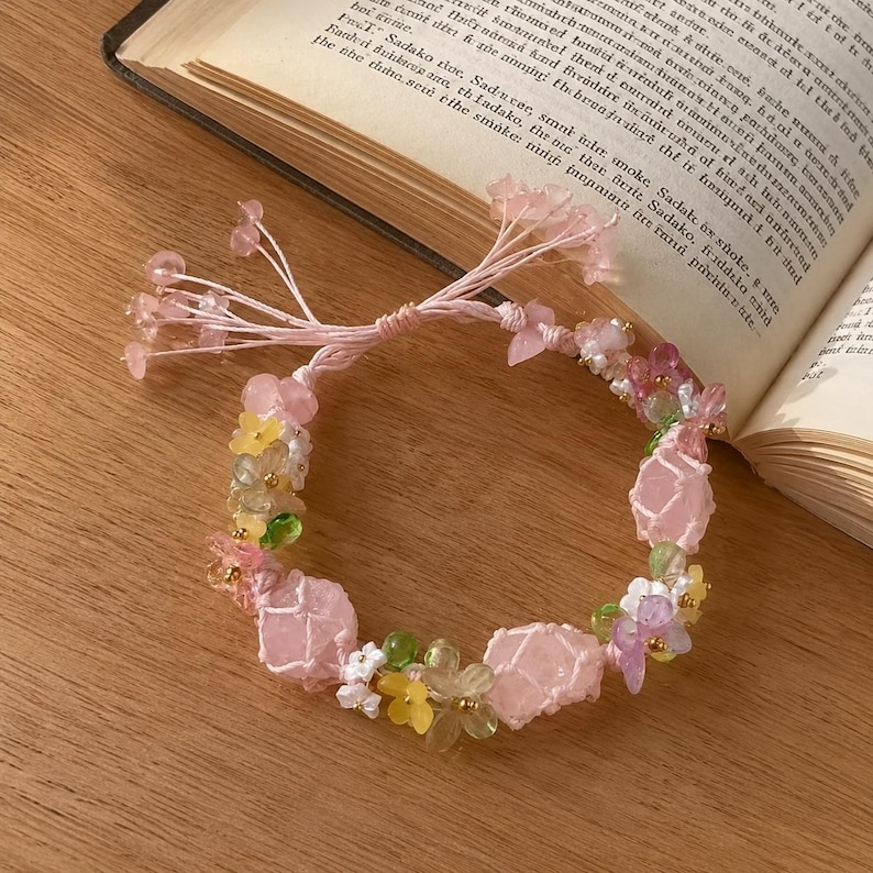 May include: A handmade bracelet featuring pink rose quartz stones, small yellow and white flower beads, and green beads. The bracelet is strung on a pink cord and is displayed on a wooden surface next to an open book.