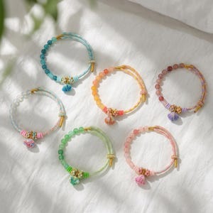 May include: A collection of seven beaded bracelets in various colors, including blue, orange, pink, and green. Each bracelet features a decorative tassel and gold-colored accents. The bracelets are displayed on a white surface.
