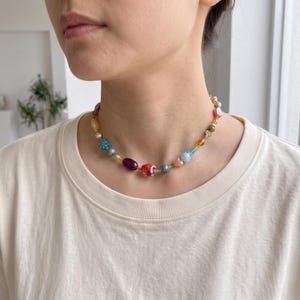 May include: A beaded necklace featuring a variety of colorful, multi-shaped beads including a small red elephant charm, translucent blue spheres, a deep purple oval bead, and various smaller accent beads in shades of pink, yellow, and green.
