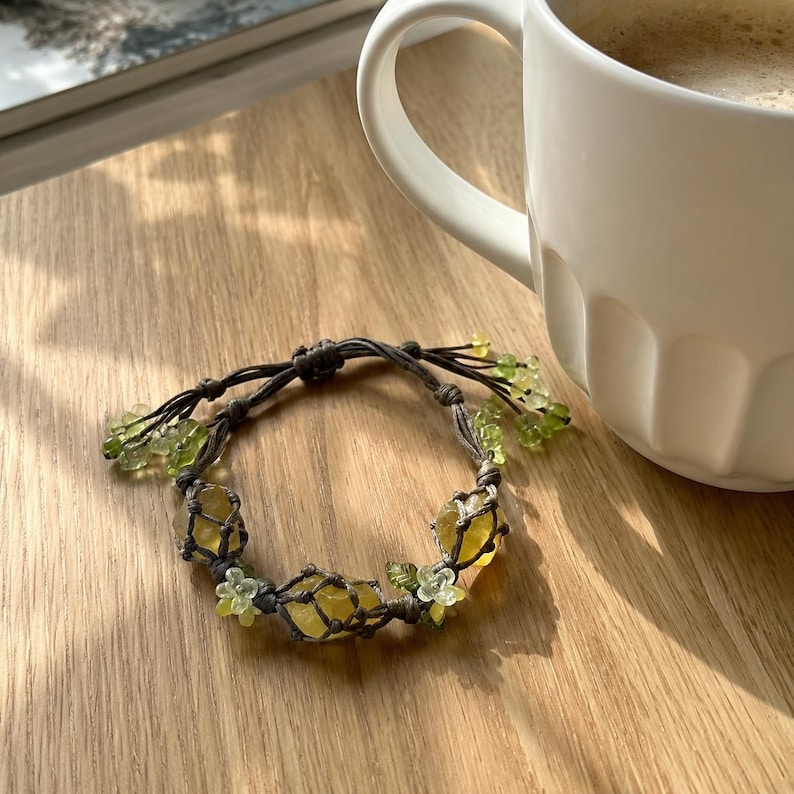 May include: A handmade bracelet featuring yellow and green gemstones, accented with small green floral beads. The bracelet is crafted with a dark gray cord and rests on a wooden surface, next to a white mug.