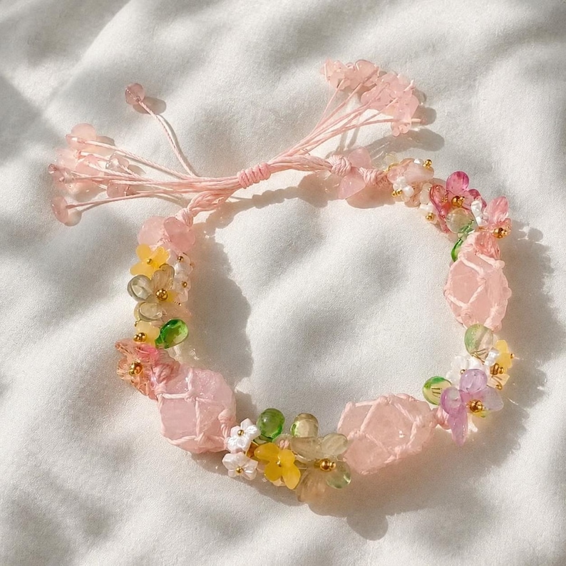 May include: A delicate pink bracelet featuring a floral design. The bracelet incorporates pink quartz stones, small colorful beads, and clusters of tiny pink flowers. The bracelet is tied with a pink cord.