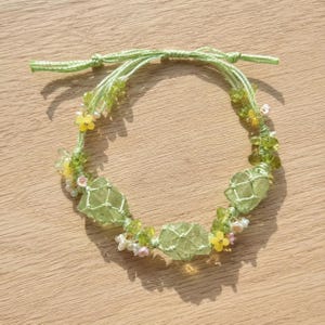 May include: A handmade bracelet featuring light green crystals encased in a macrame net design. The bracelet is adorned with small yellow and pink floral accents, and tiny white beads, all strung on a light green cord.
