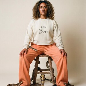 Op de afbeelding: Een persoon draagt een crèmekleurige sweatshirt met de tekst "I AM Brave" en een oranje joggingbroek, zittend op een houten kruk. Verschillende slangen zijn opgerold rond de kruk en de benen van de persoon. Neutrale achtergrond.