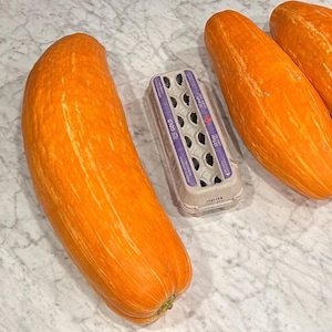 Peut inclure: Vue aérienne de plusieurs courges orange allongées avec des rayures claires, à côté d'une boîte à œufs vide. Les courges sont disposées sur une surface claire, semblable à du marbre, suggérant une cuisine ou un espace de préparation culinaire.