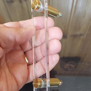 May include: Clear acrylic cabinet pull with brass fittings. The rectangular pull has a faceted edge and is attached with two brass cylinders. The hardware is a bright gold colour. The pull is held in a hand.