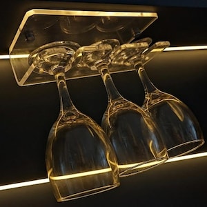 Könnte beinhalten: Ein Weinglashalter aus klarem Acryl zur Unterbaumontage mit drei Weingläsern. Der Halter ist an einer dunklen Oberfläche befestigt und verfügt über eine integrierte, warmweiße LED-Beleuchtung, die die Glasstiele und die Acrylplatte erhellt.