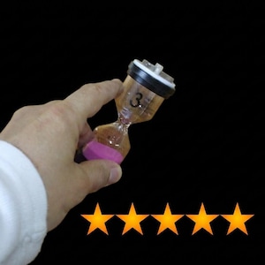 May include: A clear plastic hourglass with a black cap and a pink base, featuring the number 3 printed in black on the upper chamber. The hourglass contains a light brown granular substance and is positioned against a solid black background.