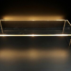 May include: A rectangular clear acrylic display riser featuring a flat top surface supported by four cylindrical metal legs at each corner. The edges of the transparent platform are illuminated with a warm light, creating a glowing effect.