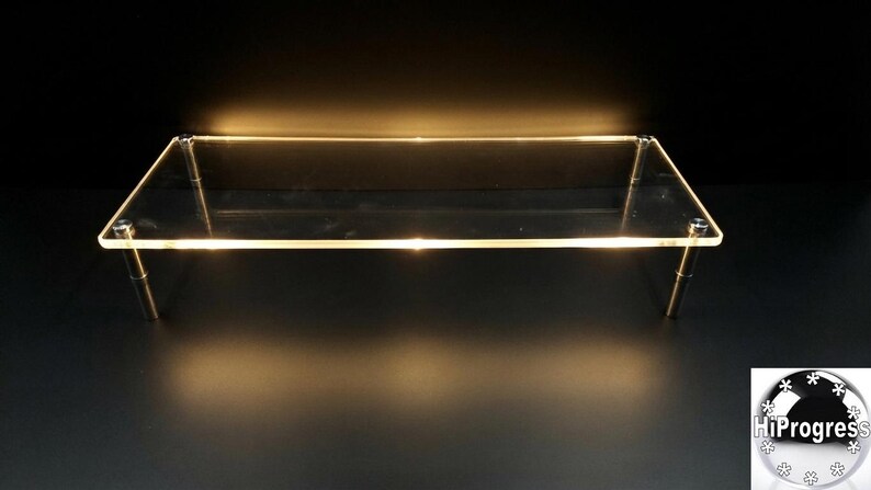 May include: A rectangular clear acrylic display riser featuring a flat top surface supported by four cylindrical metal legs at each corner. The edges of the transparent platform are illuminated with a warm light, creating a glowing effect.
