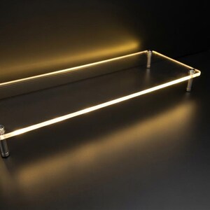 May include: A rectangular clear acrylic display riser featuring a smooth, polished surface and rounded corners. The platform is elevated by four cylindrical silver-toned metal legs positioned at each corner, creating a raised, open-frame structure.