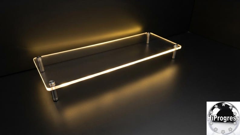 May include: A rectangular clear acrylic display riser featuring a smooth, polished surface and rounded corners. The platform is elevated by four cylindrical silver-toned metal legs positioned at each corner, creating a raised, open-frame structure.