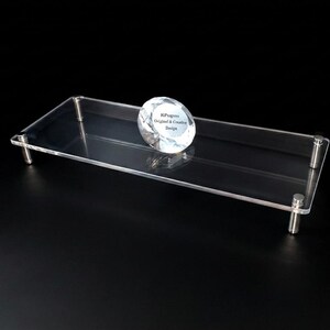 May include: A rectangular clear acrylic display riser featuring four silver-toned metal cylindrical legs. A faceted, diamond-shaped glass paperweight sits on the surface, printed with the text HiProgress Original & Creative Design.