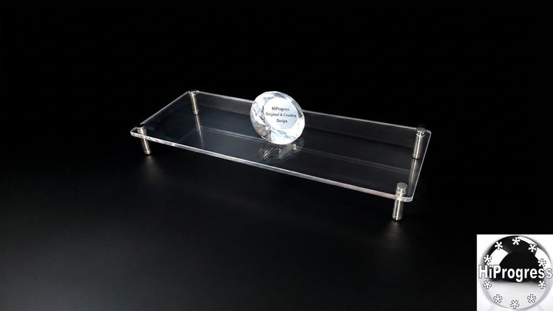 May include: A rectangular clear acrylic display riser featuring four silver-toned metal cylindrical legs. A faceted, diamond-shaped glass paperweight sits on the surface, printed with the text HiProgress Original & Creative Design.